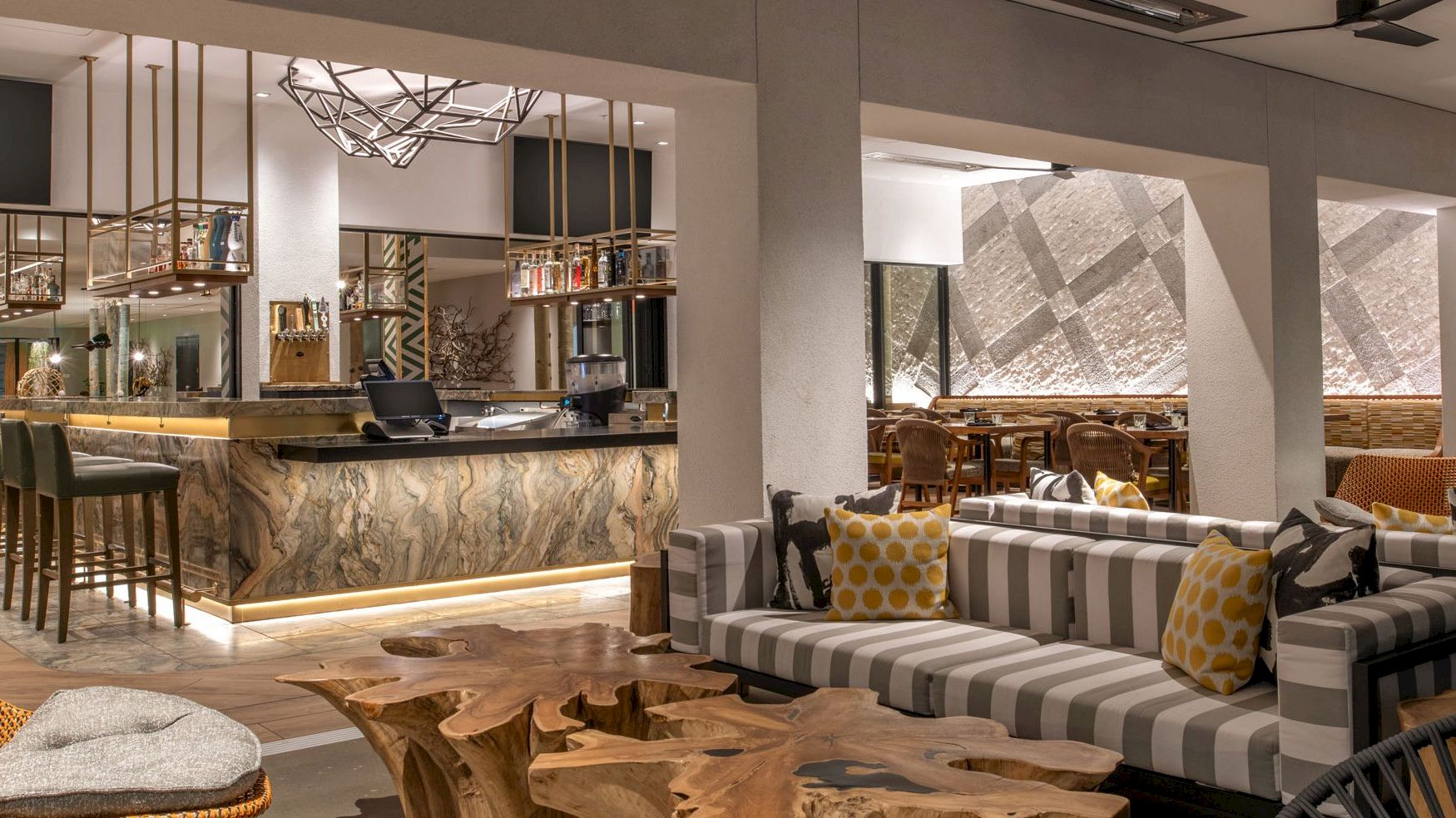 A modern, stylish lounge and bar area with a mix of seating options, including striped couches and wooden tables, and a well-lit bar in the background.
