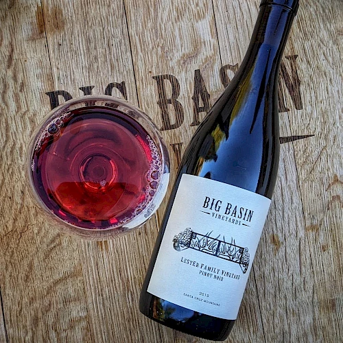 A bottle of Big Basin wine lies next to a glass of red wine on a wooden surface with "BIG BASIN" engraved.