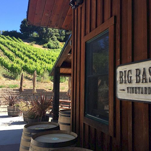 The image depicts the exterior of a winery building with a "Big Basin Vineyards" sign, wine barrels, and a vineyard on a hillside in the background.